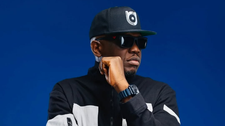 iLLBliss rests his hand near his chin in a thoughtful pose, wearing a black and white jacket, a black logo cap, and a wristwatch, set against a bold blue backdrop.