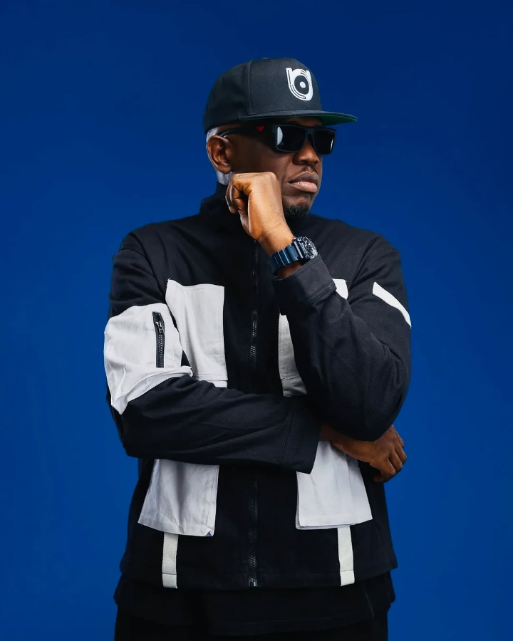 Rapper Illbliss poses with arms crossed, dressed in a black and white zip-up jacket and a black cap with a logo, standing confidently against a vibrant blue background.