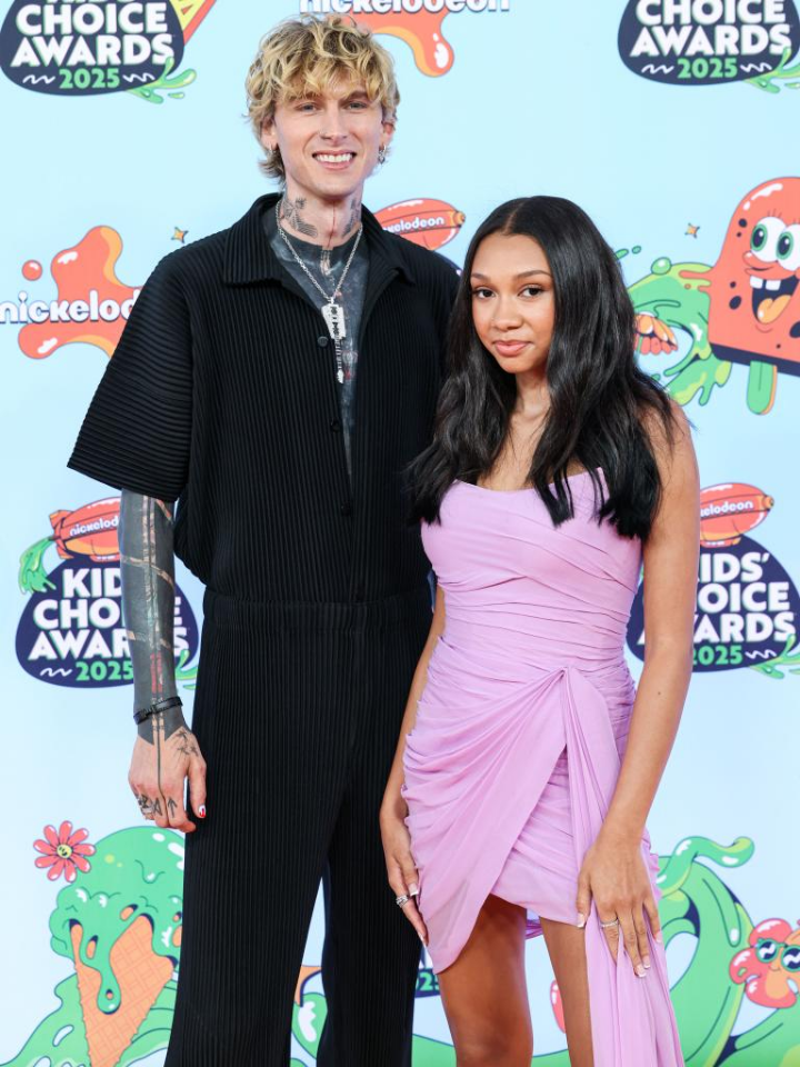 Machine Gun Kelly stands beside his daughter Casie at the 2025 Nickelodeon Kids' Choice Awards, sporting a black outfit with tattooed arms exposed, while Casie wears a pink draped dress; both smile in front of a vibrant, branded backdrop.