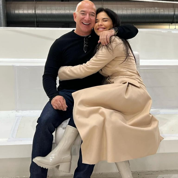 Jeff Bezos and Lauren Sanchez sit closely on a white bench, with Sanchez affectionately wrapping her arms around Bezos. He wears a black shirt and jeans, while she’s styled in a beige coat and light boots, set against an industrial backdrop.