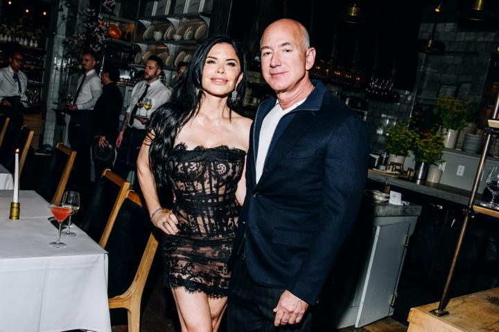 Jeff Bezos and Lauren Sanchez pose together at a stylish restaurant gathering. He wears a dark suit with a white shirt, while she stuns in a black lace dress, her hand resting confidently on her hip as they stand side by side.