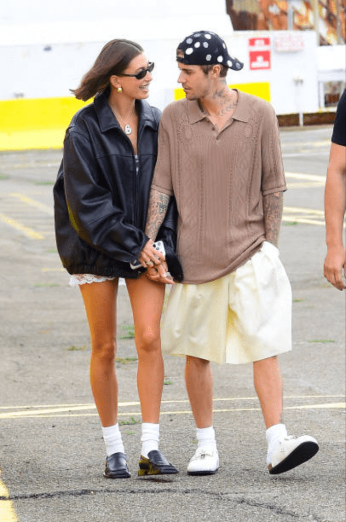 Justin Bieber and Hailey Bieber walk hand in hand during a casual outing, with Justin in a brown knit polo and white shorts, and Hailey in an oversized black leather jacket paired with white lace shorts and black loafers.