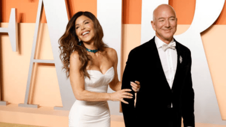 Jeff Bezos, in a classic black tuxedo, stands beside Lauren Sanchez, who wears a strapless white gown and turquoise necklace, as they pose together at an event with a bold "AIR" backdrop.
