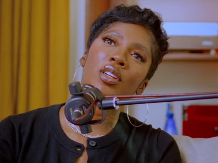 Tiwa Savage speaks into a microphone, dressed in a black top and large hoop earrings, set against a backdrop of a yellow curtain and a shelf adorned with various items.