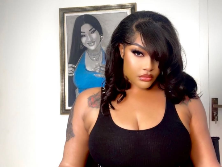 Angela Okorie stands indoors wearing a black sleeveless top, revealing tattoos on both arms, with a framed portrait of a woman in a blue top and necklace hanging on the white wall behind her.