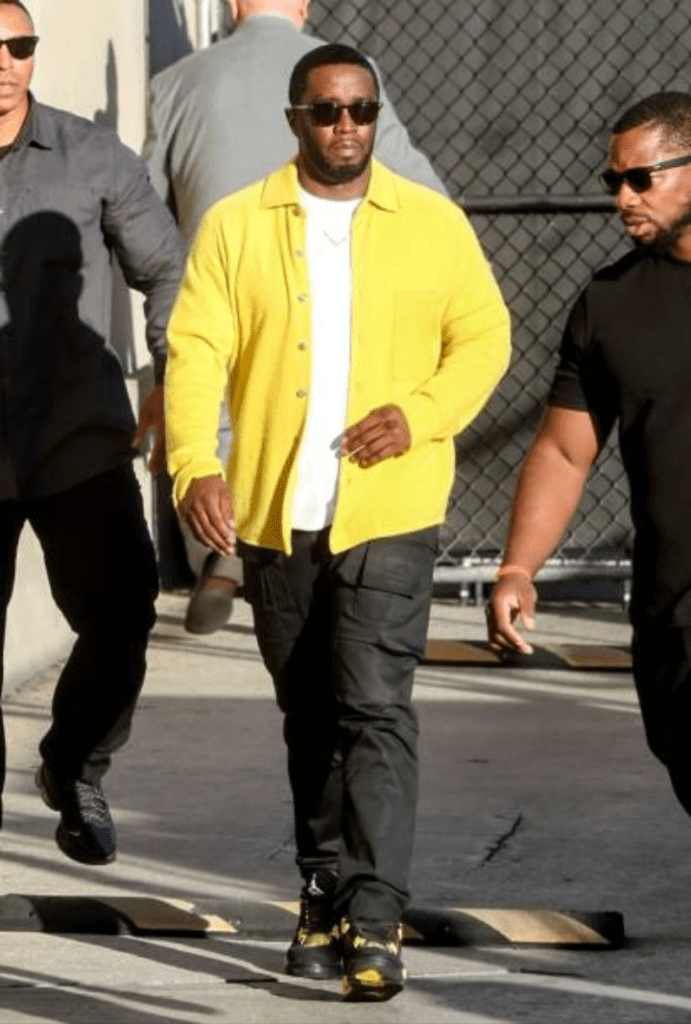 Sean “Diddy” Combs walks outdoors in a vibrant yellow jacket layered over a white shirt, paired with black cargo pants and matching sneakers featuring yellow accents. He’s flanked by two companions, with an urban backdrop of a chain-link fence and pavement adding to the streetwear vibe.