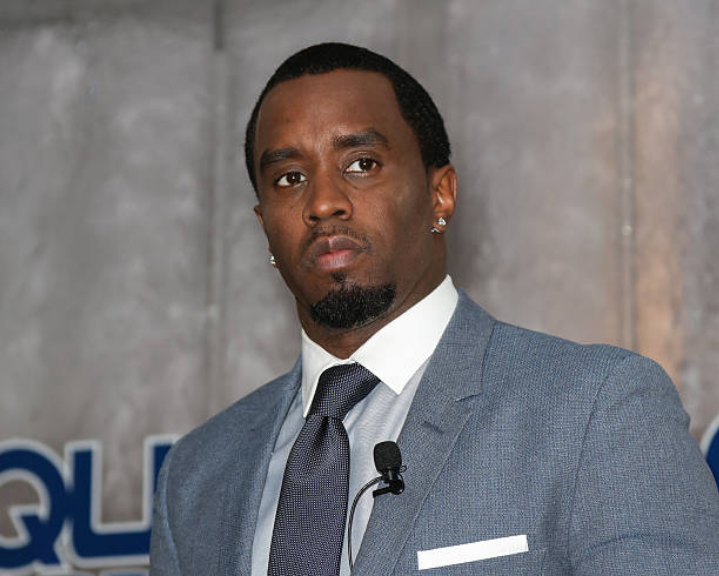 Sean “Diddy” Combs wears a light blue suit with a white dress shirt and a dark tie, accented by a microphone clipped to his tie and a stud earring. He poses against a textured wall featuring partially visible text, adding a sharp and polished aesthetic.
