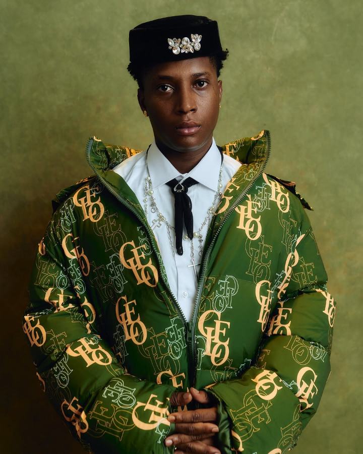 Bella Shmurda wears a green jacket with a gold "GEO" pattern, paired with a white shirt, black tie, and cross necklace, topped with a black hat featuring floral accents against a muted green background.