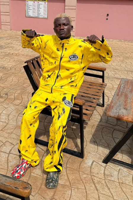 Portable sits outdoors on a wooden chair, dressed in a bold yellow tracksuit with black splatter patterns and "POLUX" printed across the jacket and pants, paired with mismatched shoes—one red and white, the other green camo—against a backdrop of a pink wall and wooden table.