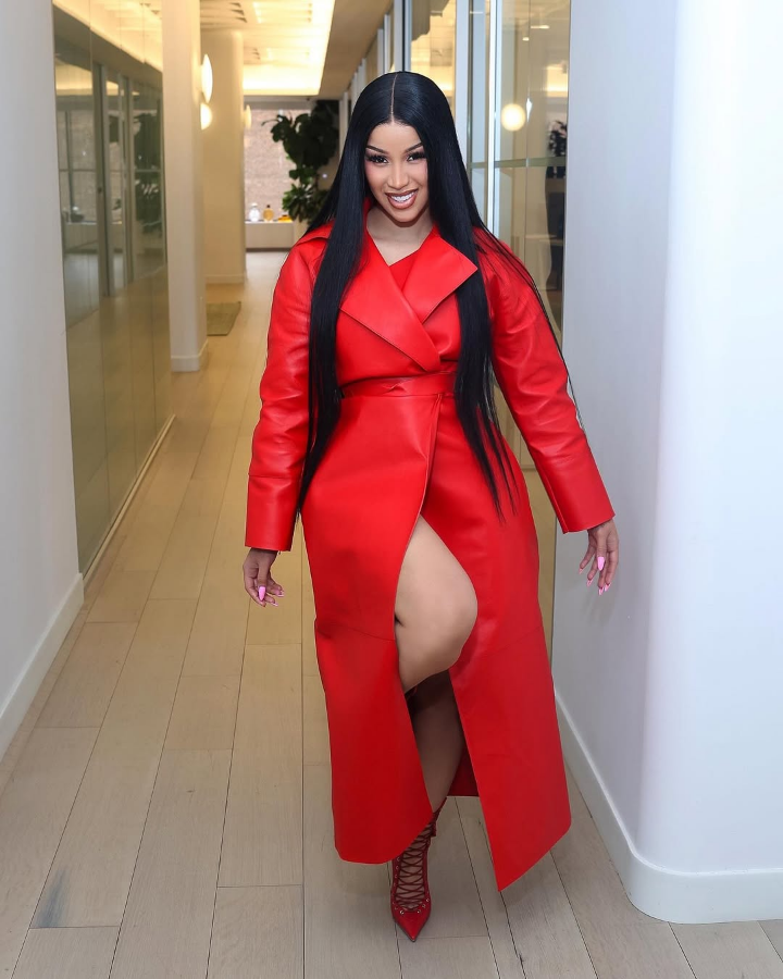 Cardi B struts down a sleek hallway in a bold red leather trench coat with a thigh-high slit, paired with matching red lace-up heels, exuding confidence and high-fashion glamour.