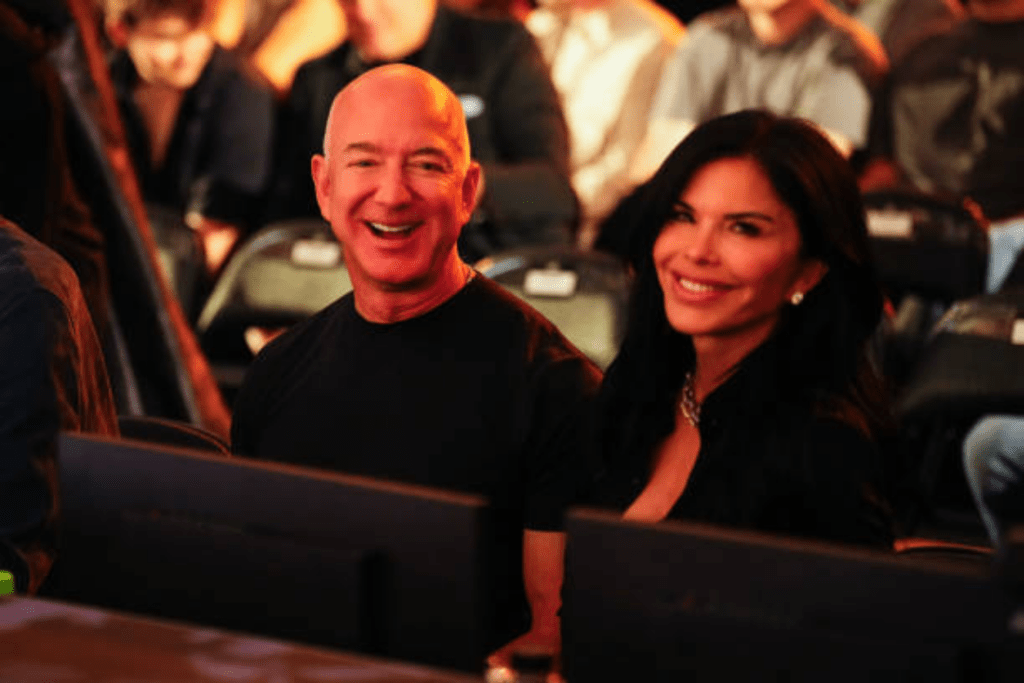 Jeff Bezos and Lauren Sanchez sit side by side at a public event, with Bezos in a black shirt and Sanchez wearing a black outfit accented by a necklace, both framed by monitors and surrounded by other attendees.