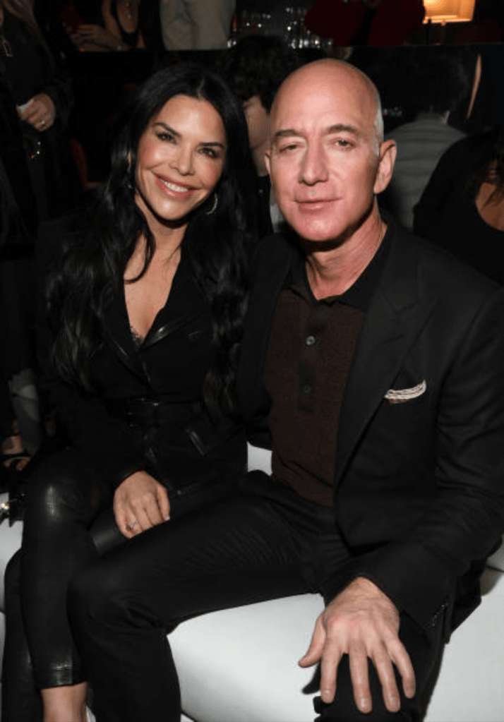 Jeff Bezos and Lauren Sanchez sit closely together at a lively event, with Bezos in a black suit and brown shirt, and Sanchez in a sleek black outfit, both appearing relaxed and engaged in the moment.
