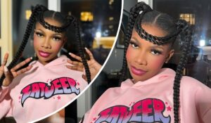 BBNaija star Tacha poses in a pink hoodie emblazoned with “ZATWEER” in vibrant lettering, accented by small stars. Her hair is styled in two long braids, which she holds confidently with both hands, against a backdrop of city lights glowing through a window.