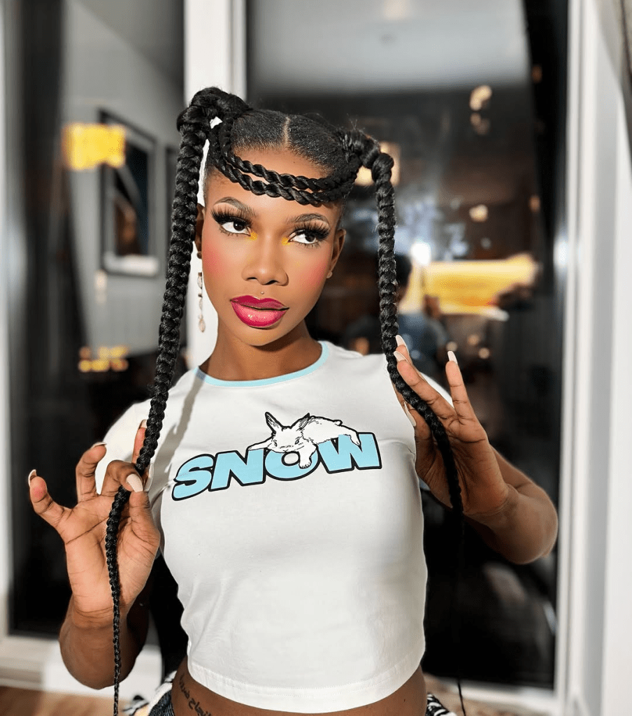 BBNaija star Tacha stands confidently in a white crop top adorned with a bold blue "SNOW" print and a unicorn graphic, holding her twin braids against a softly lit, windowed backdrop.