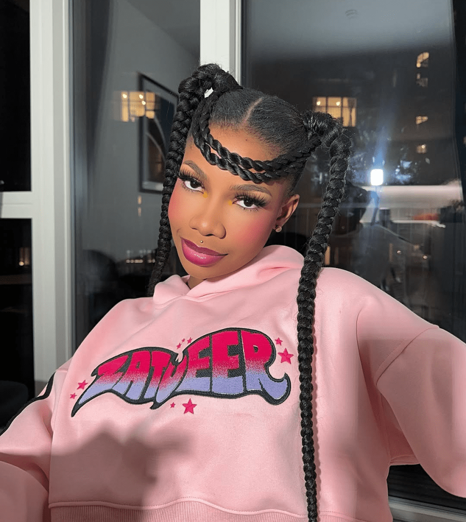 BBNaija star Tacha wears a pink hoodie with “ZATWEER” printed in a colorful gradient and star accents. She gazes confidently ahead while holding her long braids, set against a backdrop of glowing city lights through tall windows.