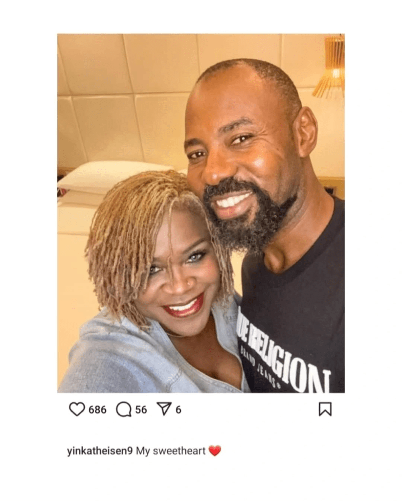 Linc Edochie poses beside his estranged wife Yinka in a warmly lit bedroom, with Yinka in a light-colored top and Linc wearing a black “True Religion Brand Jeans” t-shirt. A beige padded wall and neatly arranged white pillows form the cozy backdrop.