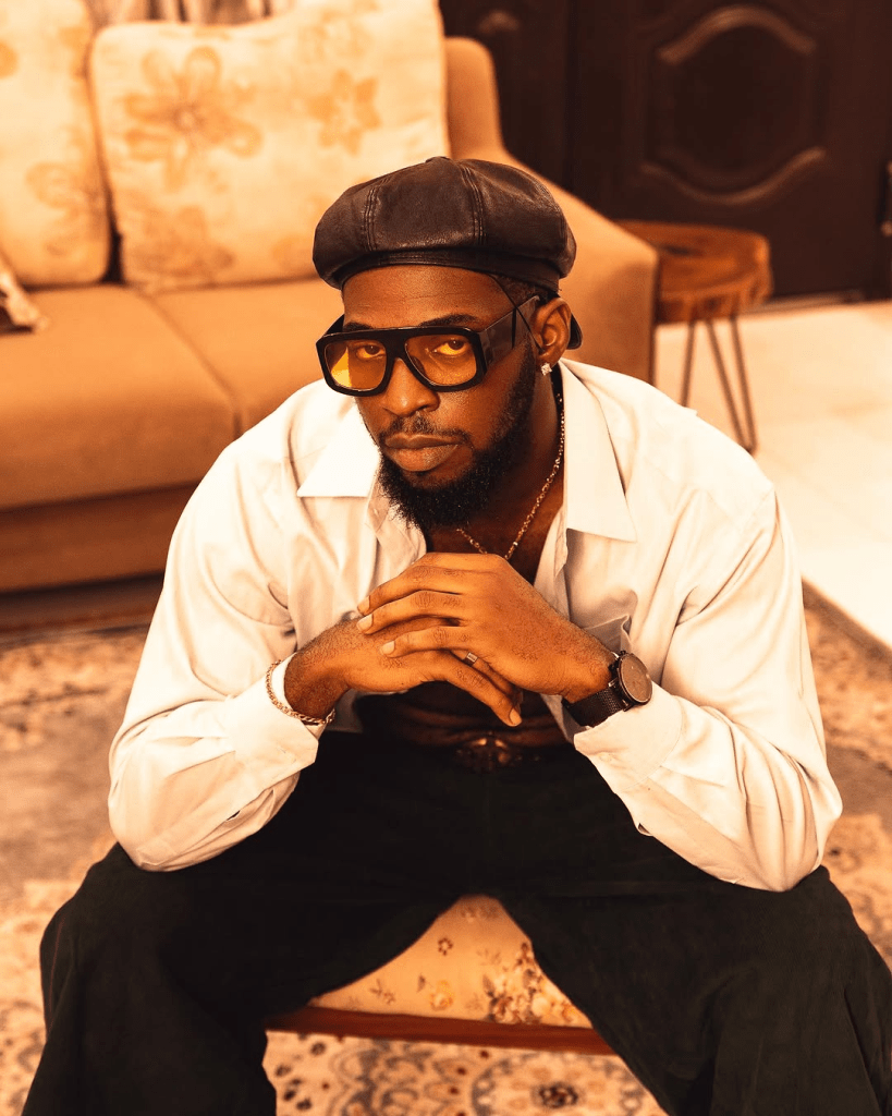 BBNaija star Zion sits on a patterned stool in a warmly lit living room, dressed in a crisp white shirt, dark trousers, and a cap. With hands clasped and a calm expression, Zion exudes a relaxed confidence amidst floral cushions and soft-toned decor.