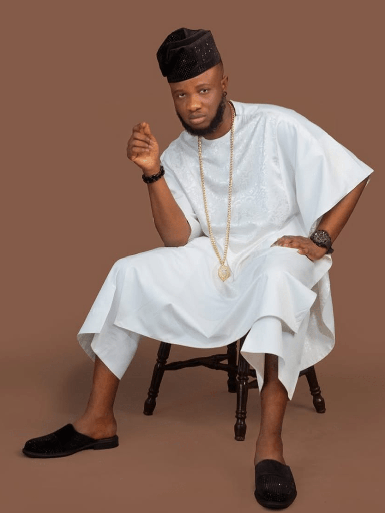 Deeone sits on a wooden chair, dressed in a white agbada with intricate embroidery. He completes the look with black slip-on shoes, a matching black cap, a gold chain, and accessories that add a modern flair against the warm brown backdrop.