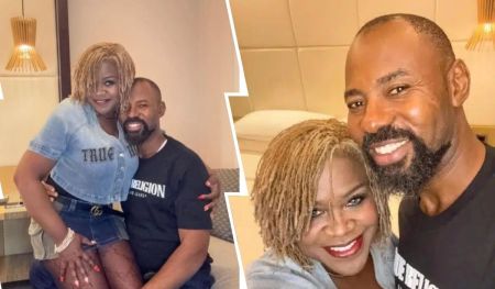 Actor Linc Edochie sits relaxed in a casual black "True Religion" t-shirt as Yinka, his estranged wife, stands close beside him with her hand resting gently on his shoulder. She wears a denim top and matching skirt with fishnet stockings, both branded "True Religion." The setting is an intimate room with warm lighting, a bed in the background, and a wooden ceiling lamp overhead.