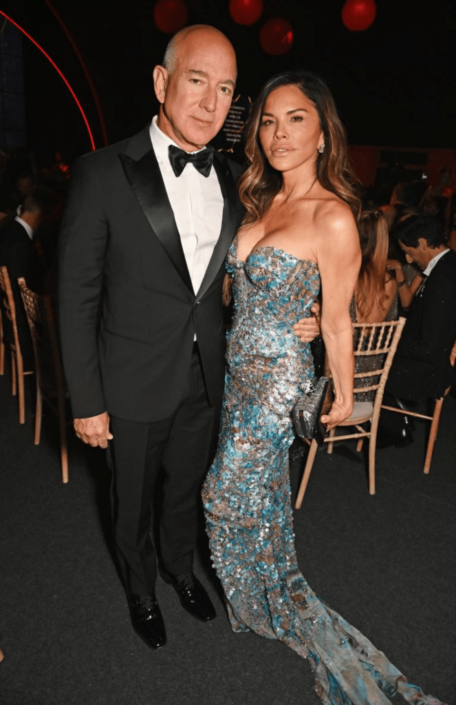 Jeff Bezos, in a classic black tuxedo, stands beside Lauren Sanchez, who wears a shimmering sequin gown with a flowing train. They appear poised for a formal evening, with Sanchez holding a clutch and elegant lighting in the background.