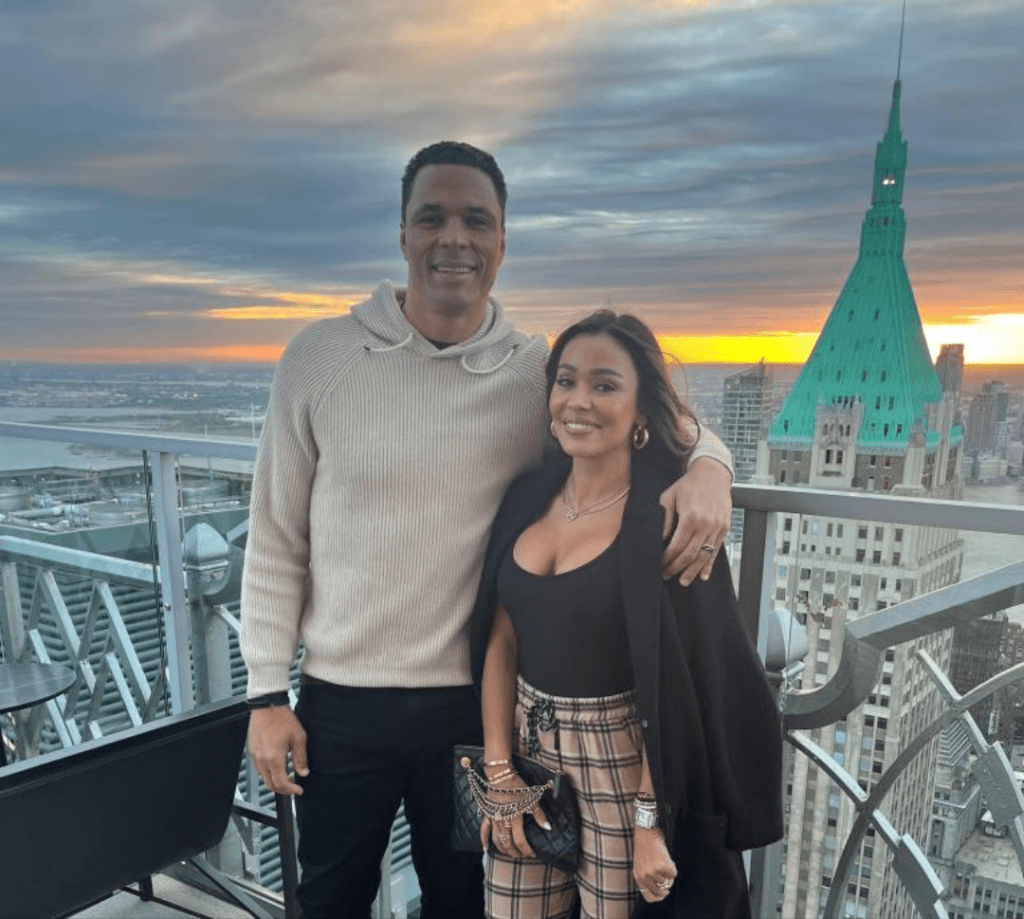Tony Gonzalez and his wife Tobie pose side by side on a rooftop at sunset, with a glowing city skyline behind them. Tony wears a light sweater, while Tobie is dressed in a black top and plaid pants, holding a clutch. A dramatic sky and a green-roofed building add visual charm to the background.