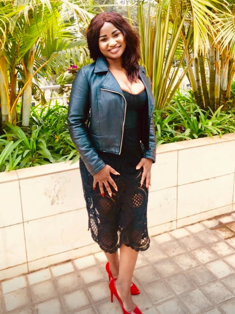 Helen Atti stands confidently on a tiled walkway, framed by vibrant tropical plants, wearing a black lace-accented dress and a sleek leather jacket, complemented by bold red heels.
