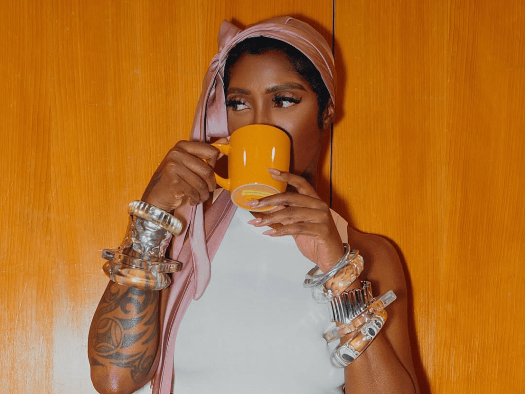 Tiwa Savage wears a white sleeveless top and a pink headscarf, accessorized with clear and silver bracelets. She holds a small cosmetic container with both hands, seated against a wooden background. A tattoo is visible on her left forearm.