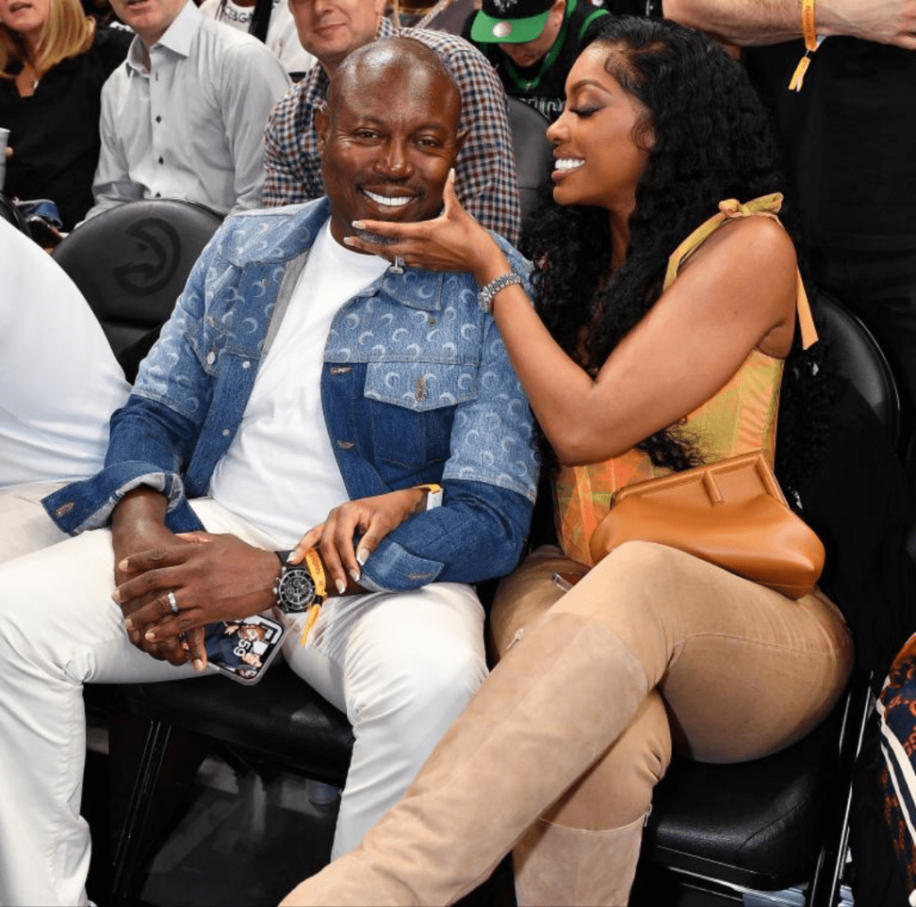 Seated at a stylish event, Simon Guobadia pairs a denim jacket with crisp white trousers, while Porsha Williams complements him in a colorful top, beige pants, and knee-high boots. She gently touches his face with one hand, her phone in the other, as they share a moment in a lively crowd.