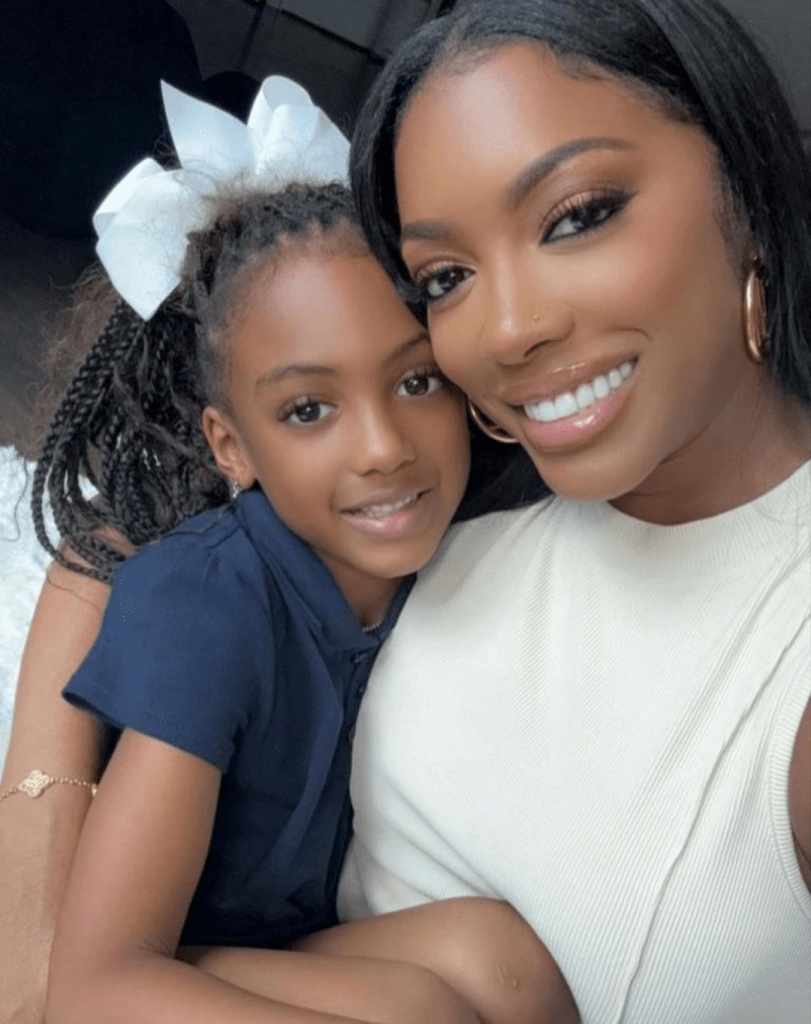 Porsha Williams cradles her daughter, who is dressed in a navy outfit with braided hair adorned by a prominent white bow. Porsha wears a sleeveless white top, accessorized with gold hoop earrings and a bracelet, as they share a warm, affectionate moment.