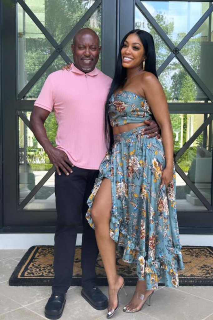 Simon Guobadia wraps an arm around Porsha Williams as they pose outside a grand glass-paneled entrance. He sports a light pink polo with black trousers, while she stands out in a floral crop top and high-low skirt, paired with clear heels. Lush greenery peeks through the backdrop, adding a touch of elegance to the stylish moment.