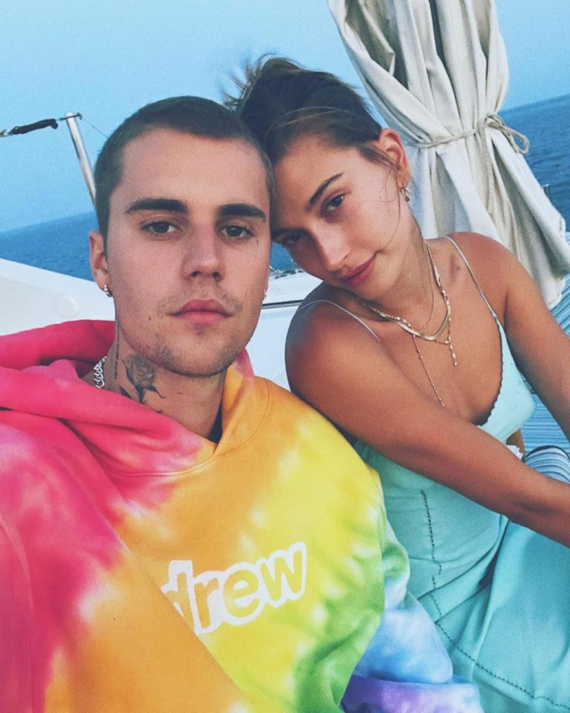 Justin Bieber wears a colorful tie-dye hoodie with "drew" on the front, while Hailey Bieber sits beside him in a light blue dress with delicate straps and layered necklaces. They appear relaxed and close, seated on a boat with the ocean stretching out behind them.