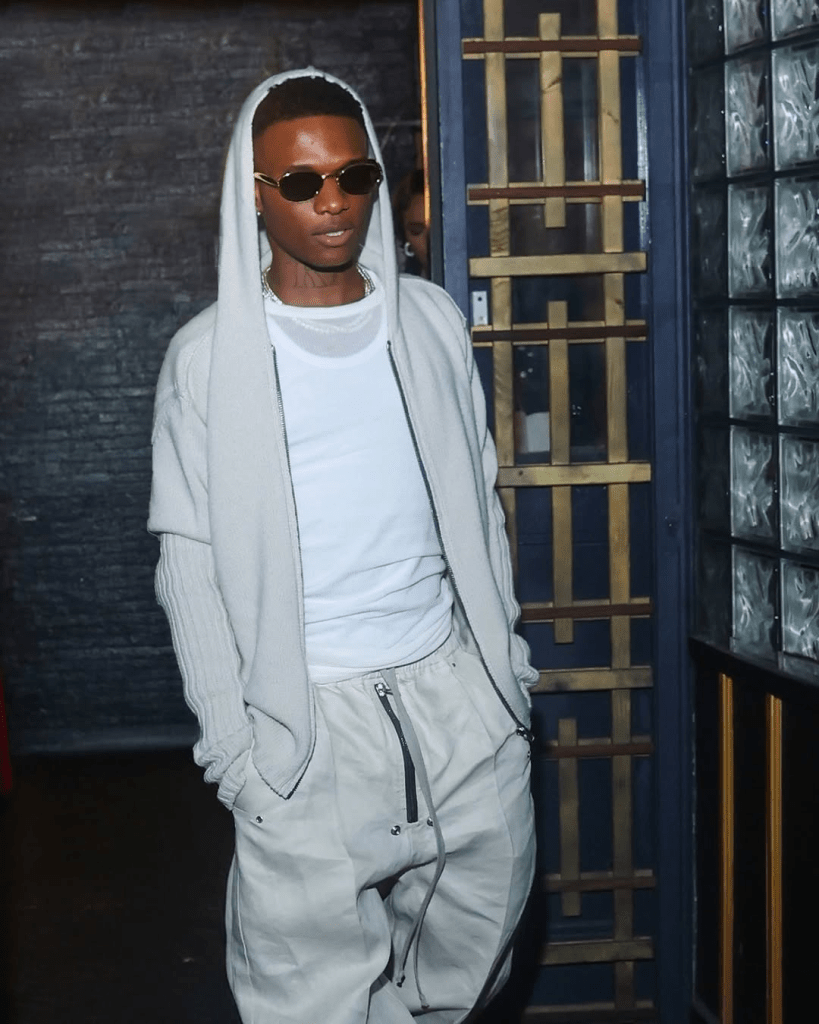 Wizkid stands in front of a dark brick wall, dressed in a white t-shirt layered under a light gray hooded cardigan and beige pants with zipper details. His relaxed pose and neutral-toned outfit reflect a cool, understated style.