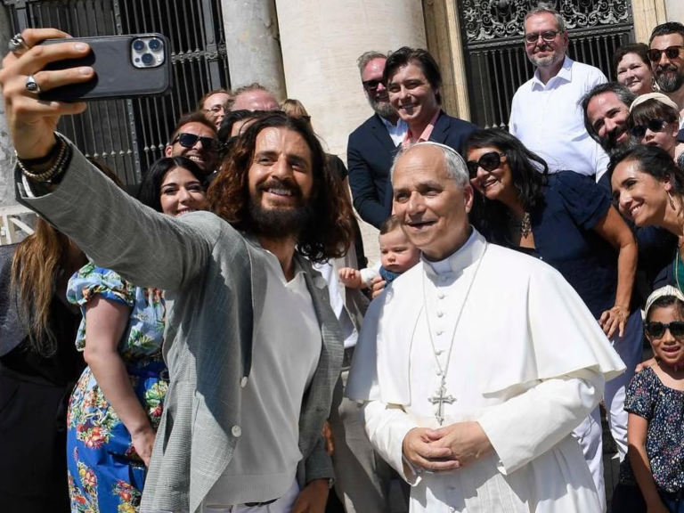 Pope Leo XIV, in traditional white papal attire, stands beside actor Jonathan Roumie, who is capturing the moment with a selfie. They are joined by a cheerful crowd near an ornate building entrance, blending faith and popular culture in a candid encounter.