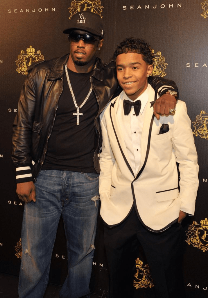 Diddy, in a black leather jacket, matching cap, and cross necklace, stands confidently beside his son Justin, who wears a sharp white tuxedo with black accents and a bow tie. The pair pose against a branded Sean John backdrop featuring a gold crest.