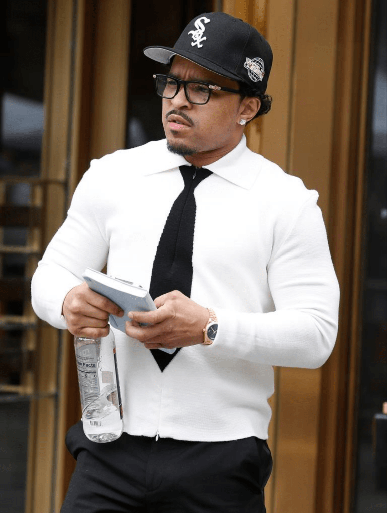 Justin Combs wears a white sweater layered over a black tie, styled with black pants and a Chicago White Sox cap. He holds a water bottle and a tablet, accessorized with a gold wristwatch.