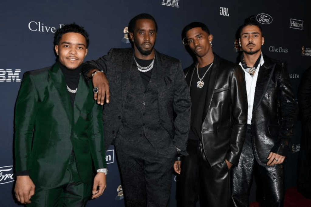Diddy poses alongside his three sons, all dressed in distinctive formalwear. They stand confidently against a backdrop featuring sponsors like Clive Davis and Hilton. Their outfits include a green suit, a black patterned ensemble, a leather jacket, and a glossy black suit, complemented by bold accessories and polished shoes.