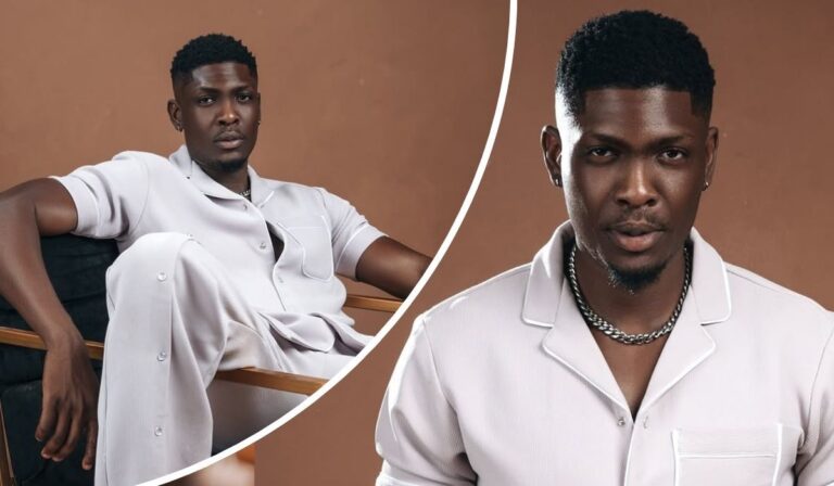 Seated in a wooden chair with a black cushion, BBNaija star Sammie exudes relaxed confidence in a coordinated light-toned outfit—short-sleeved button-up shirt, matching pants, and a silver chain necklace—set against a warm, earthy backdrop that enhances the stylish composition.