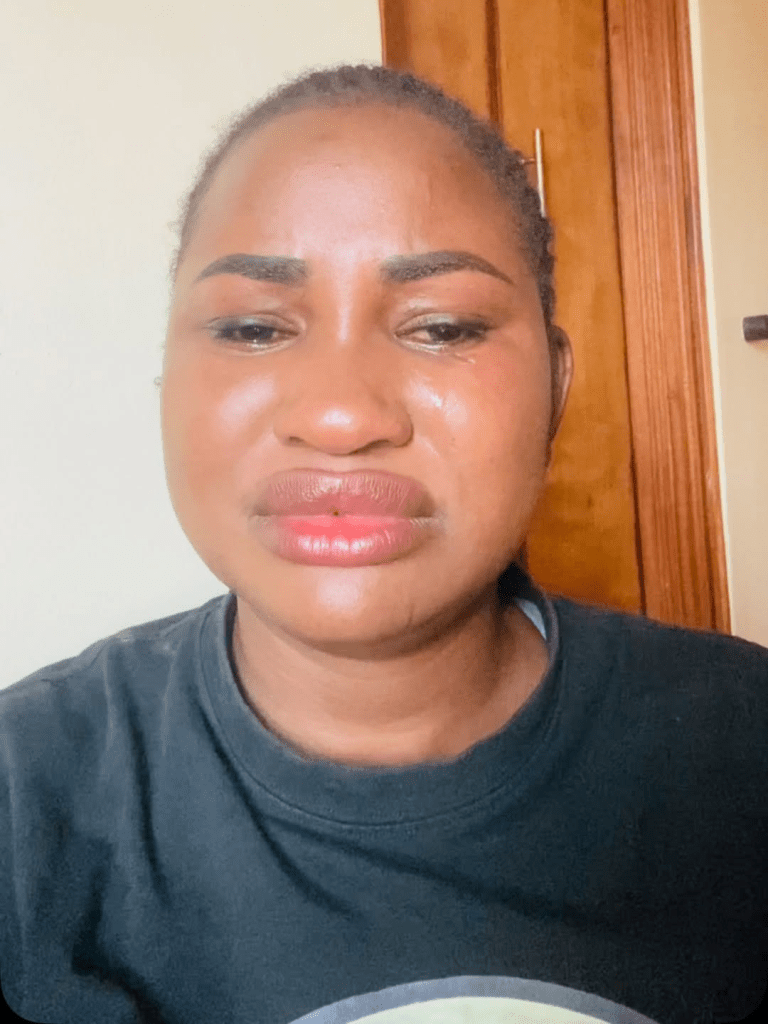 Hellen Ati poses against a light-colored wall near a wooden door, wearing a black t-shirt with a bold circular graphic. Her relaxed stance and confident expression add a casual-chic vibe to the scene.