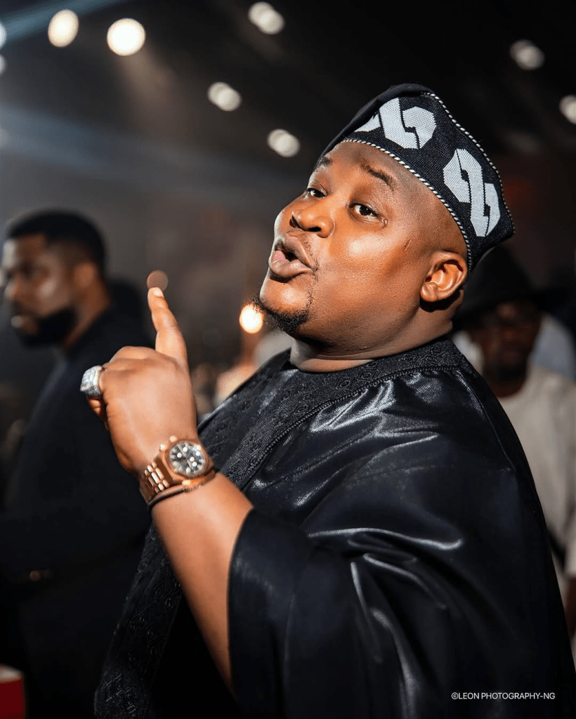 Cubana Chief Priest stands confidently in a black agbada adorned with intricate embroidery, complemented by a matching cap. He gestures expressively with one hand, showcasing a gold wristwatch and a large ring, while vibrant event lighting and fellow guests fill the background. Photo credit: LEON PHOTOGRAPHY-NG.