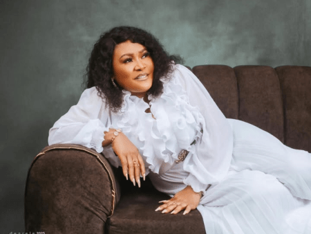 Mummy Dolarz reclines gracefully on a brown sofa, dressed in an elegant white ensemble with dramatic ruffles. Her look is polished with manicured nails, sparkling accessories, and a serene expression, set against a soft grey backdrop that adds to the sophisticated ambiance.