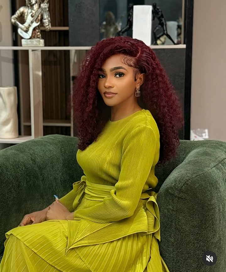 Kassia sits on a green couch in a striking yellow pleated outfit with a tied waist detail, surrounded by artistic décor including a sculpture of a musician playing a guitar, exuding elegance and creative flair.