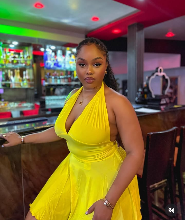 Onyeka stands confidently at a stylish bar, glowing in a halter-neck yellow dress, with colorful backlighting and sleek bottles creating a vibrant, upscale atmosphere.