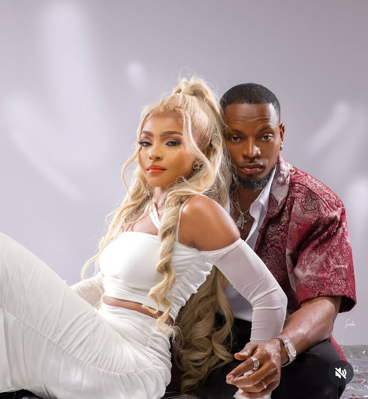 BBNaija stars Kellyrae and Kassia sit side by side in coordinated style—Kellyrae in a sleek white outfit with cut-out shoulders, and Kassia in a red patterned shirt layered over a white top, accessorized with a watch and rings against a softly blurred backdrop.