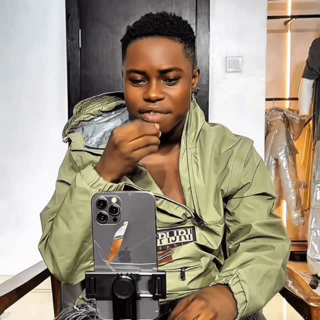 Tiktoker Peller sits in front of a smartphone on a tripod, wearing a light green Napapijri jacket, with a black door and hanging clothes in the background.