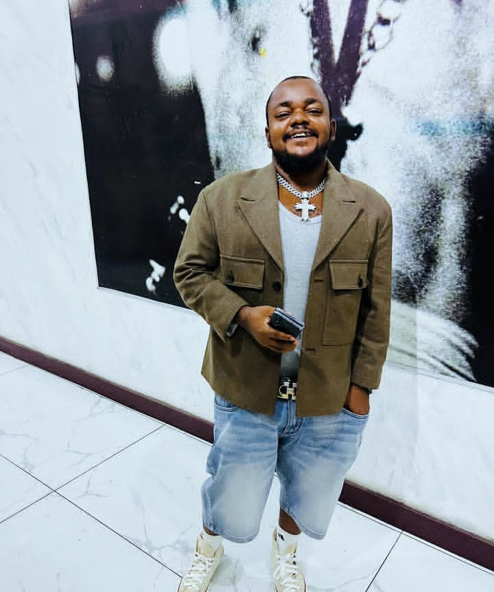 Odira Nwobu stands confidently in a brown jacket over a gray shirt, light blue denim shorts, and white sneakers, accessorized with a large cross necklace and holding a phone, set against a marble wall adorned with black and white artwork.