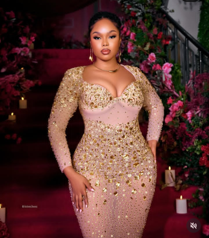 BBNaija star Onyeka poses confidently in a gold-embellished gown with a deep neckline and long sleeves, set against a lavish floral and candlelit backdrop that radiates elegance and glamour.