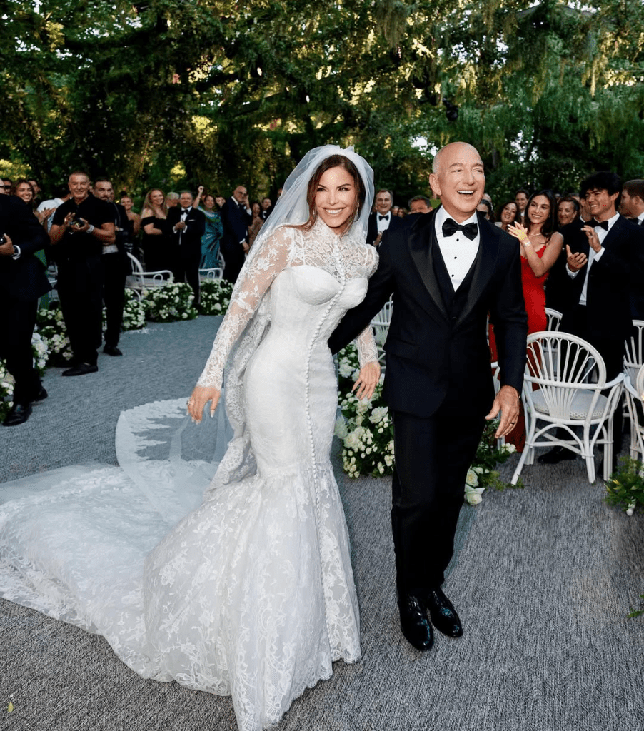 Jeff Bezos and Lauren Sanchez walk hand in hand down the aisle, beaming with joy after their wedding ceremony. He wears a classic black tuxedo with a bow tie, while she stuns in a white lace gown with a flowing train and veil. Guests cheer around them amid lush greenery and elegant floral arrangements.