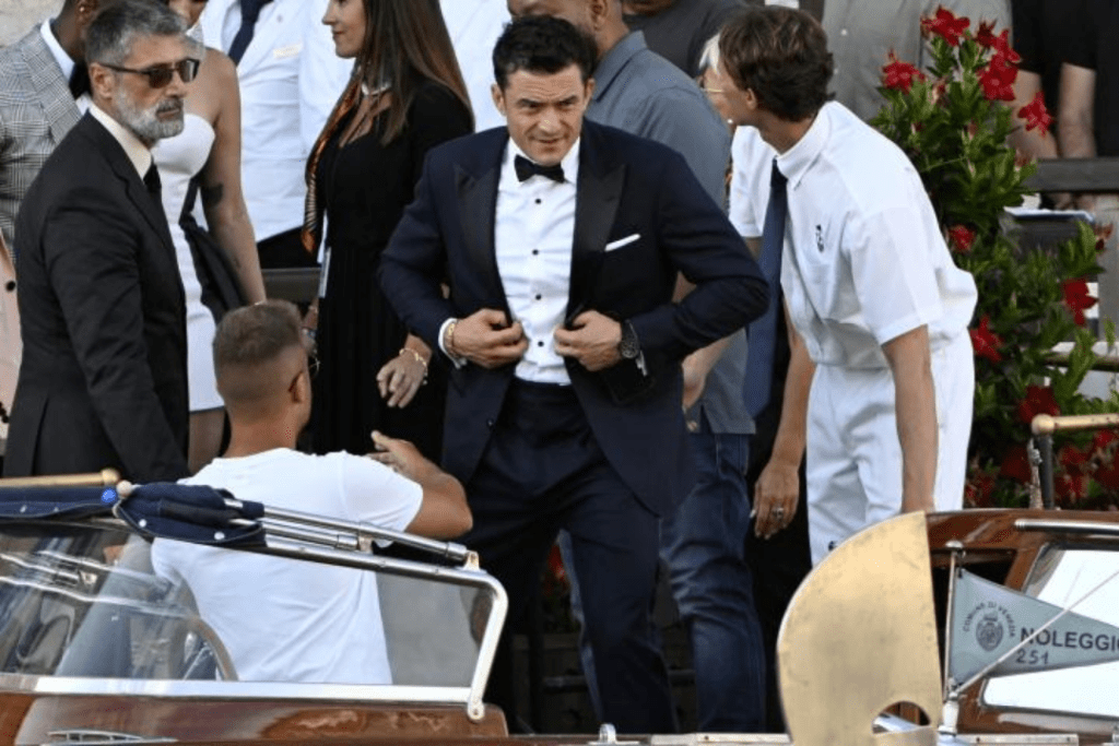 Orlando Bloom adjusts his navy blue suit jacket while standing on a dock during a formal gathering. He’s surrounded by elegantly dressed guests, with a wooden boat labeled "NOLEGGIO 251" in the foreground and vibrant red flowers in the background.