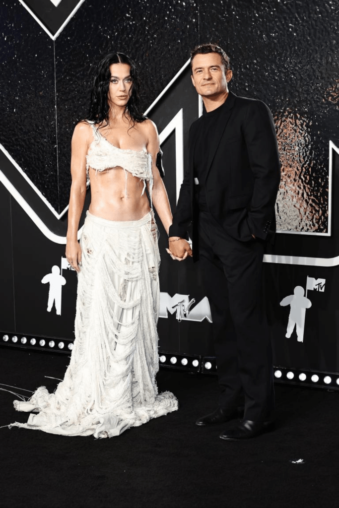 Katy Perry and Orlando Bloom hold hands at an MTV event, with Katy in a white, distressed two-piece outfit and Orlando in a sleek black suit. They pose against a backdrop featuring the MTV logo and bold geometric patterns.
