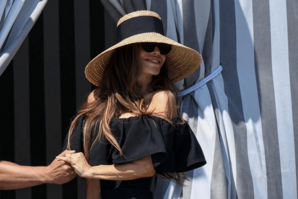 Jeff Bezos and Lauren Sanchez hold hands in front of grey-and-white striped curtains, with Sanchez wearing a wide-brimmed straw hat and a black off-the-shoulder dress.